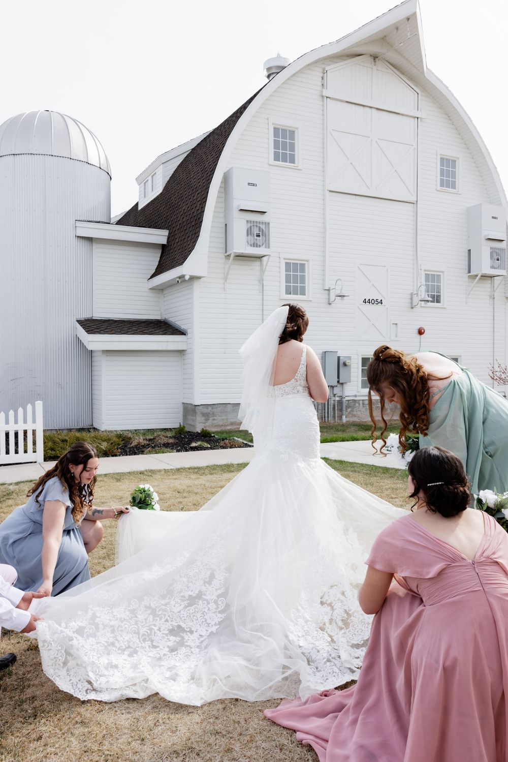 Bridesmaids in blue and pink dresses arrange the lace train of a bride in a mermaid gown outside a white barn.