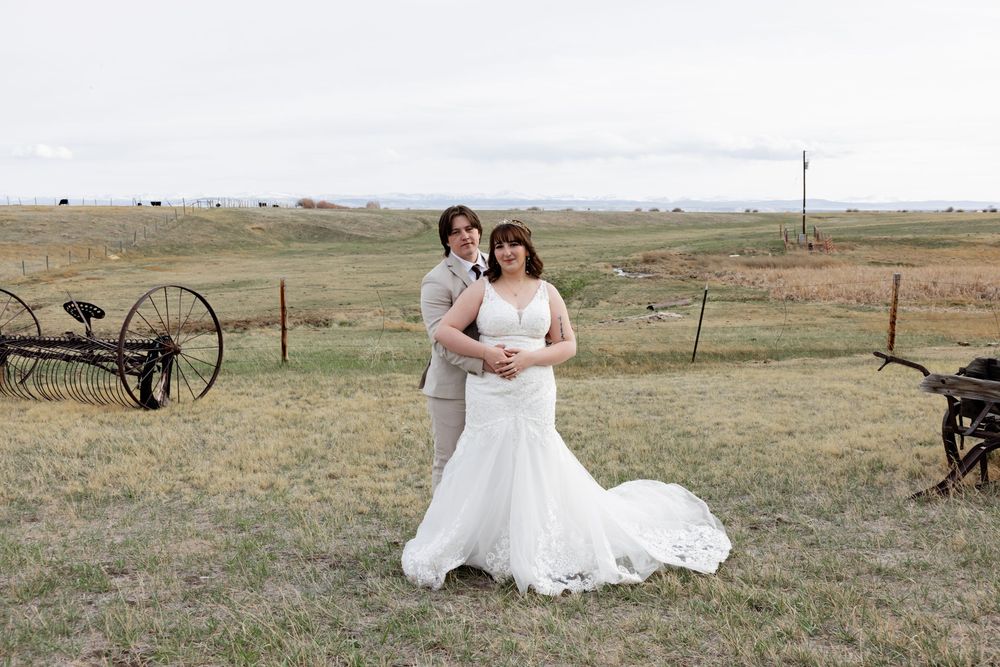 Bride in white lace mermaid gown and groom in gray suit pose in rustic farm field on their wedding day.