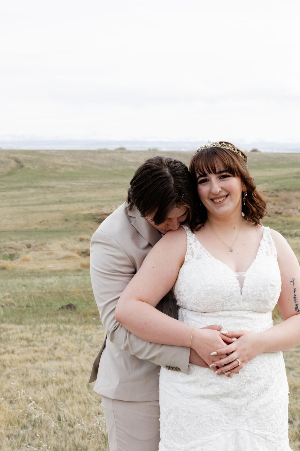 A bride in a white lace gown and tiara smiles as her partner in a tan suit embraces her in an open field.