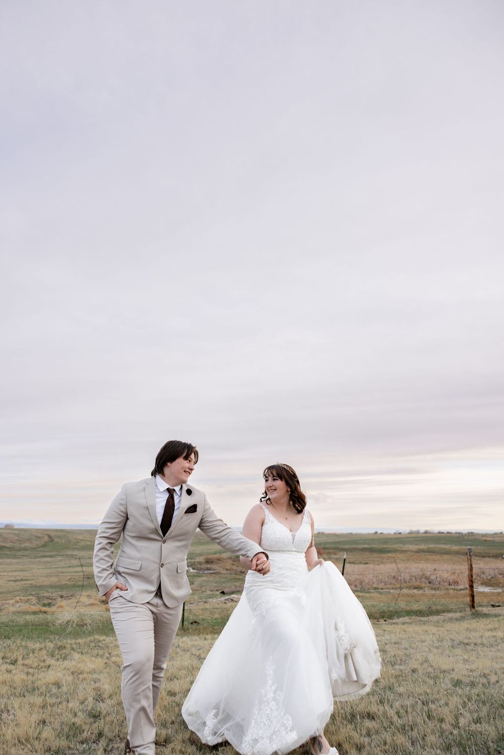 A bride and groom walk hand-in-hand through an open field at dusk, smiling at each other on their wedding day.