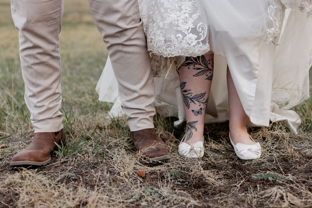Close-up of wedding couple's feet, bride showing leg tattoo in white heels, groom in tan pants and brown cowboy boots.