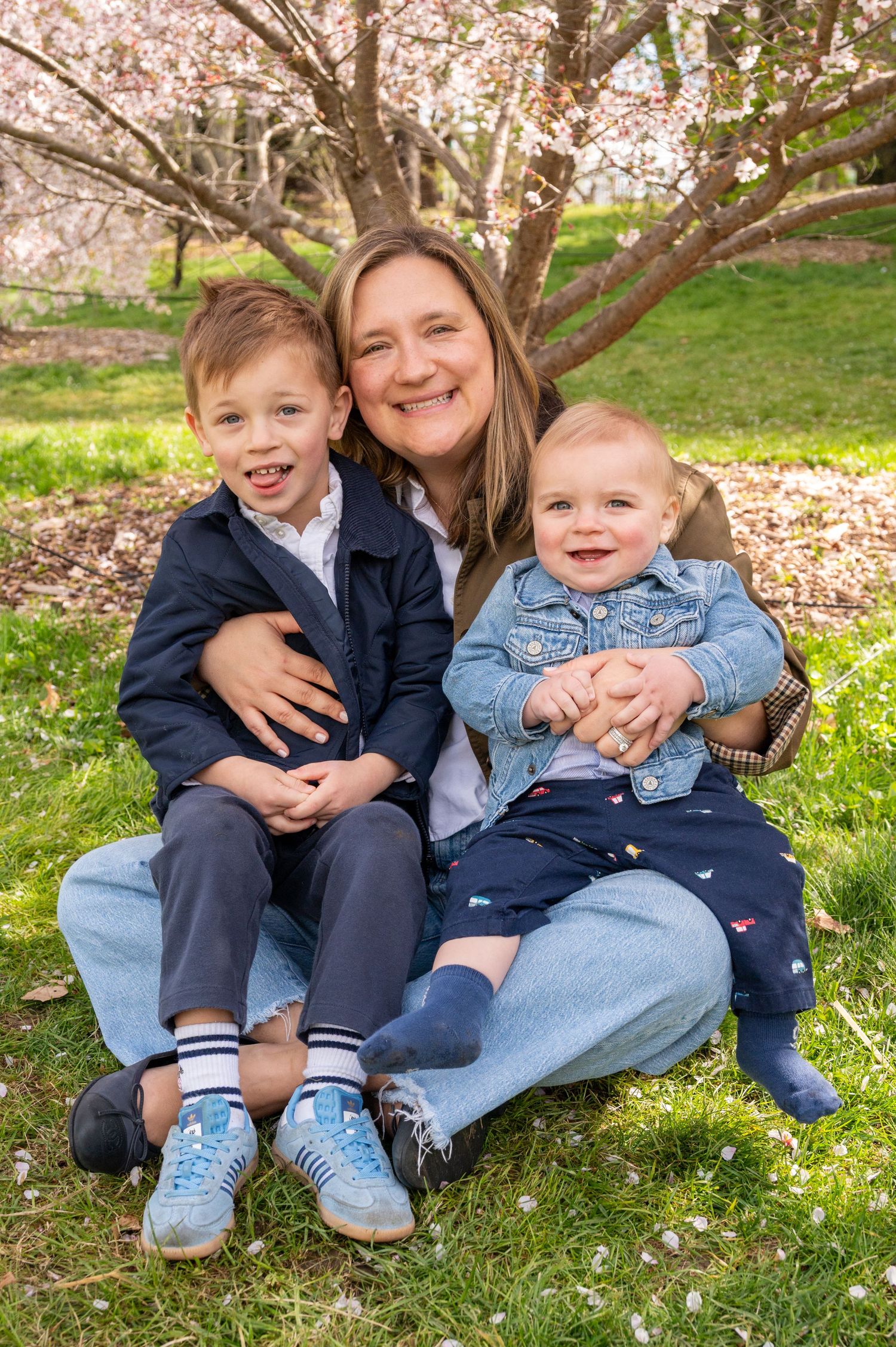 A smiling woman sits on grass under blooming cherry trees, holding two young children in a sunny spring park.