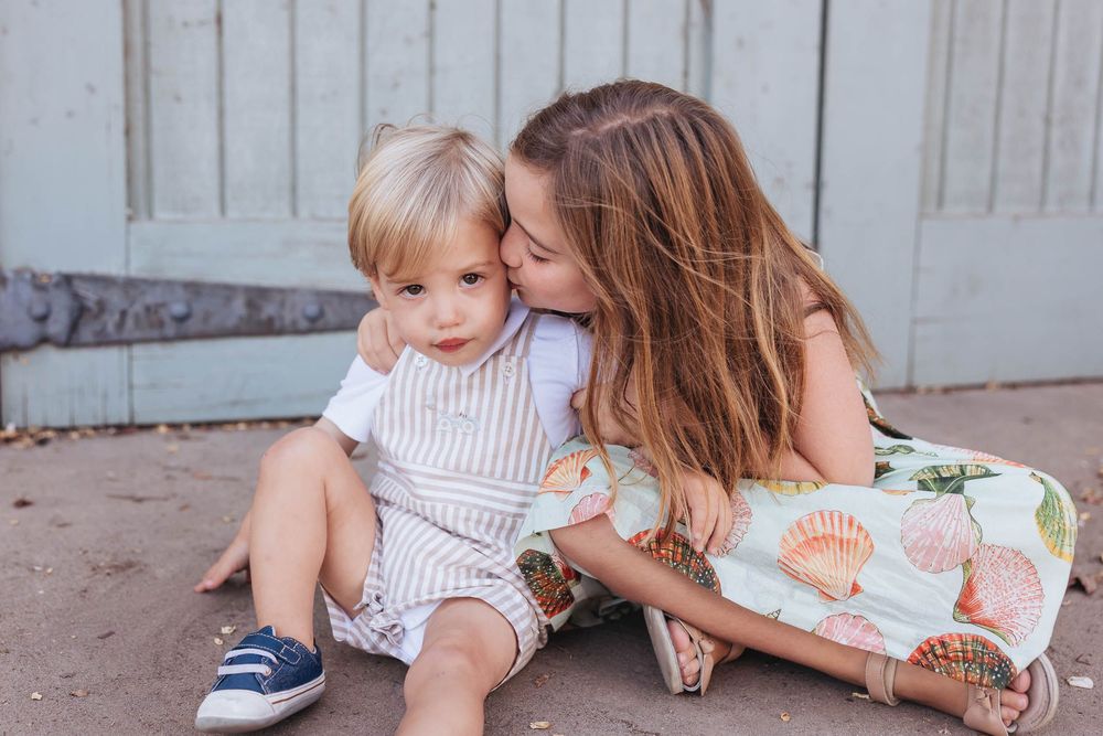 A young girl kisses a toddler boy on the cheek as they sit together outdoors near a rustic door.