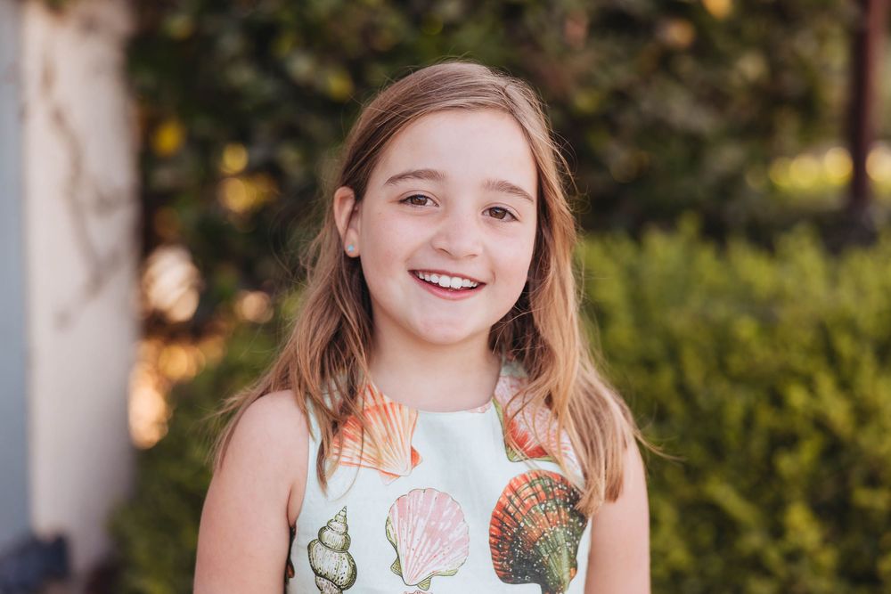 Smiling young girl with blonde hair wearing a seashell print dress outdoors with green foliage background.