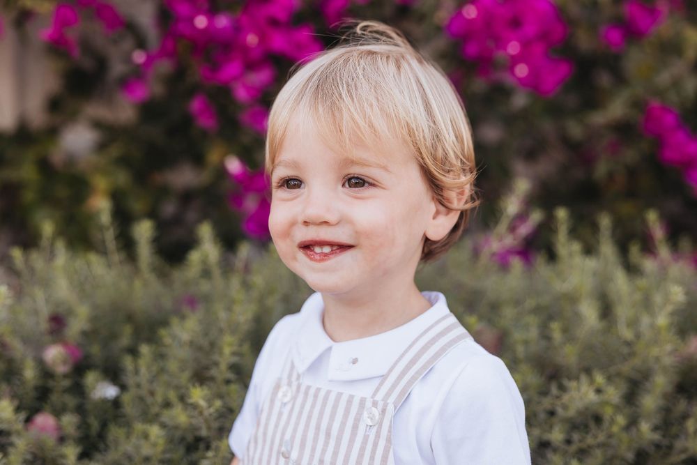 Smiling blonde toddler boy in striped overalls and white shirt with pink flowers in background.