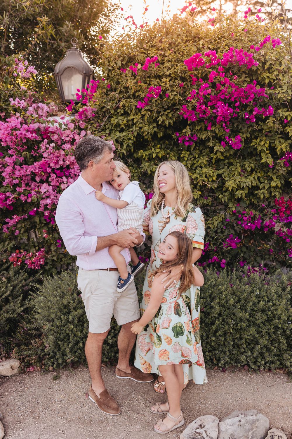 Family of four posing happily in front of vibrant pink bougainvillea, parents holding children in a sunny garden setting.