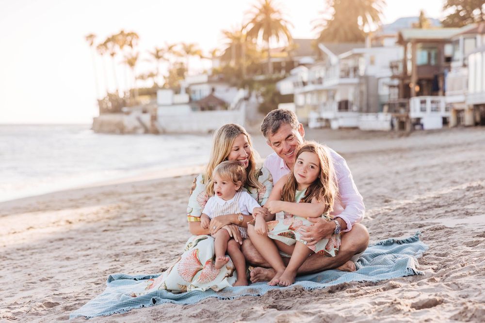 Family of four sitting on a blanket on a sunny beach, smiling and hugging with coastal homes in the background.