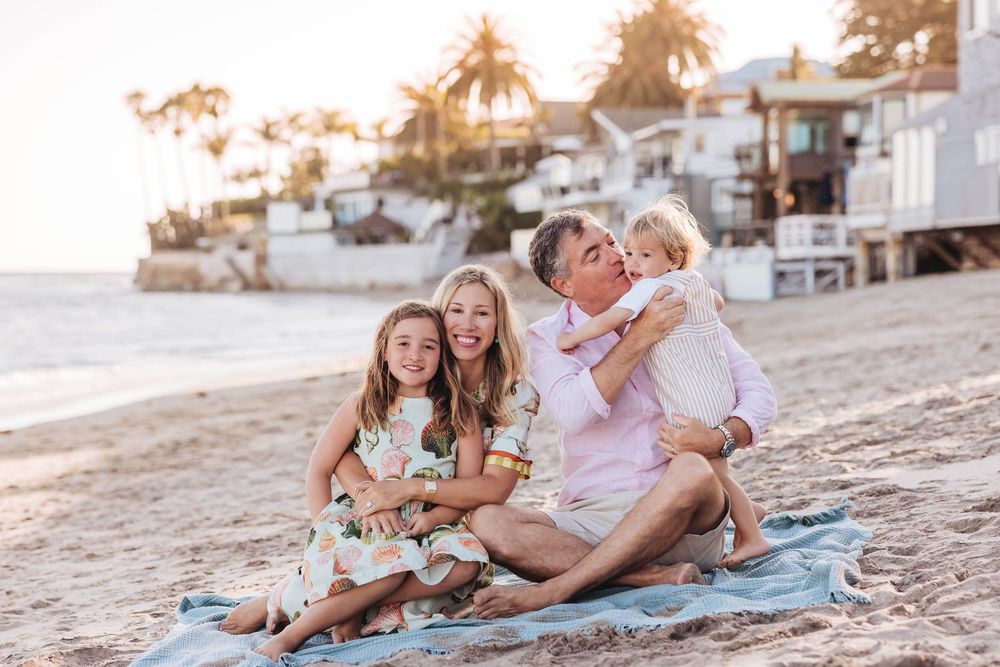 Family of four smiling together on a sandy beach at golden hour, with coastal homes and palm trees in the background.