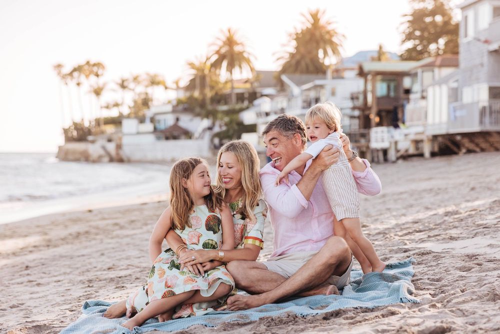 Family of five enjoying a sunny beach day, sitting on a blanket with palm trees and coastal homes in the background.