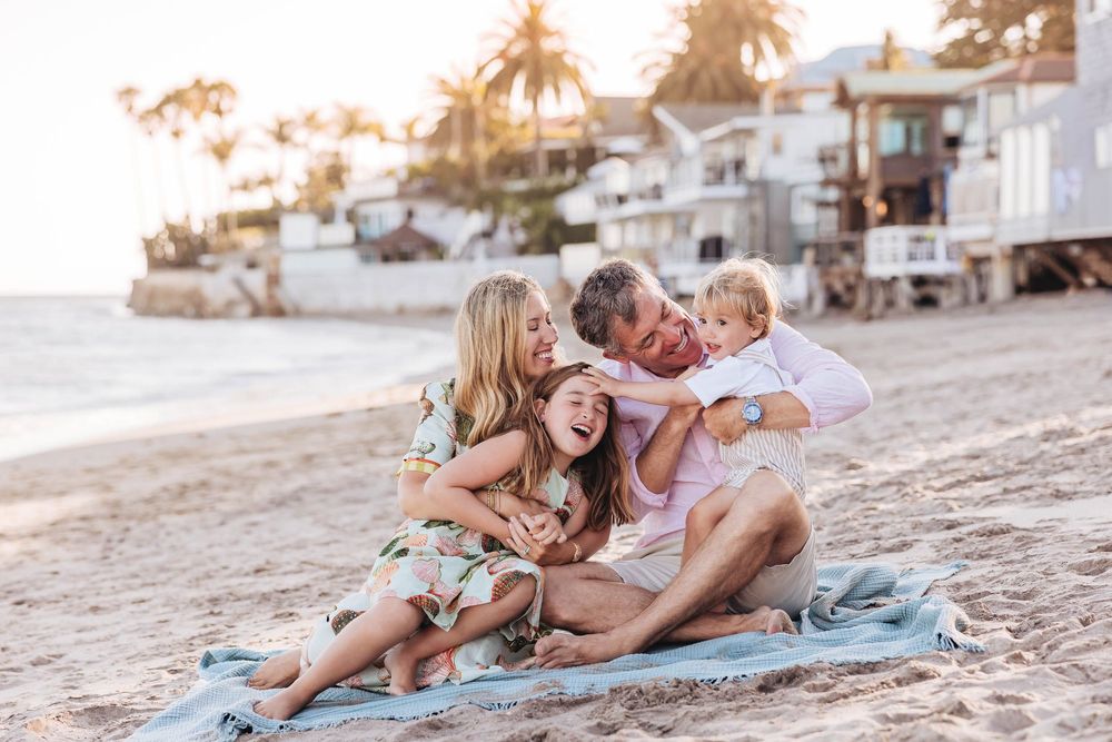 Family laughing together on sandy beach at sunset, sitting on blanket with white beach houses in background.