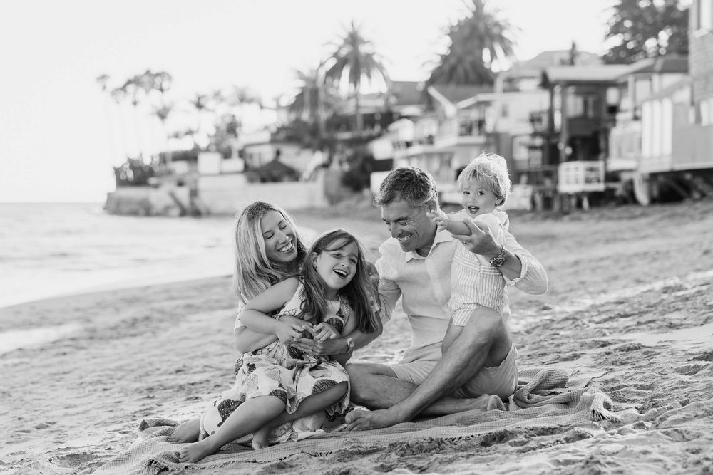 Black and white photo of a happy family of four sitting on a sandy beach, laughing and enjoying time together.
