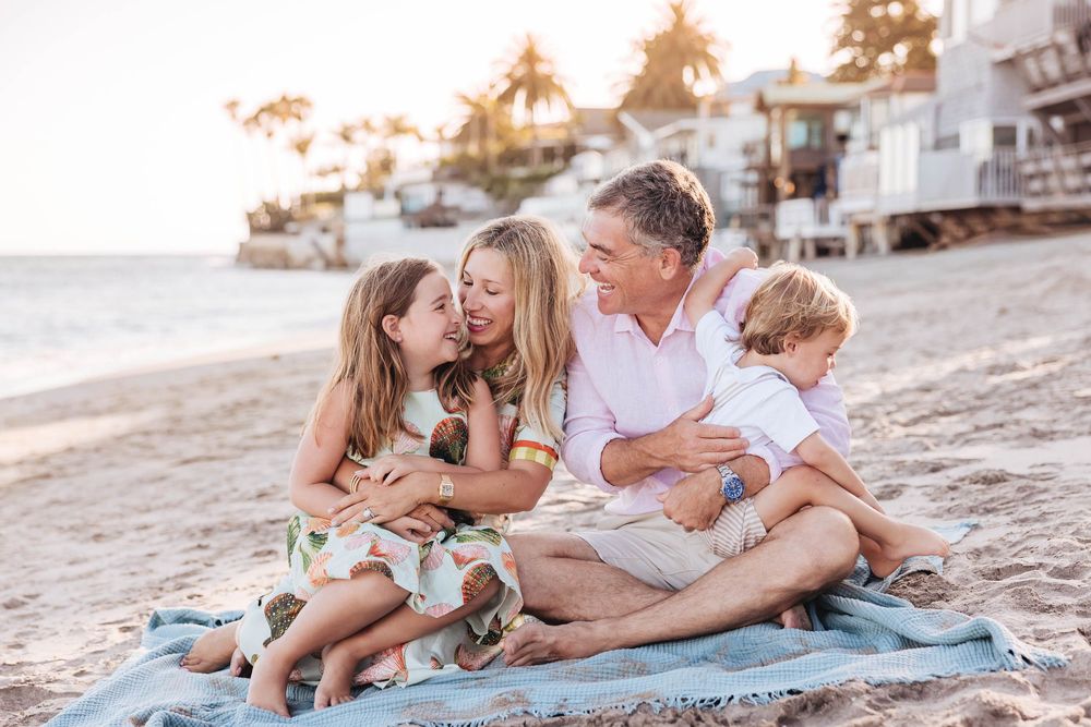 Family of four sitting on a blanket on the beach at sunset, smiling and hugging together.