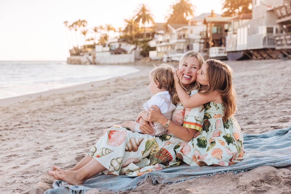 Three children share a joyful hug while sitting on a blanket on a sunny California beach.