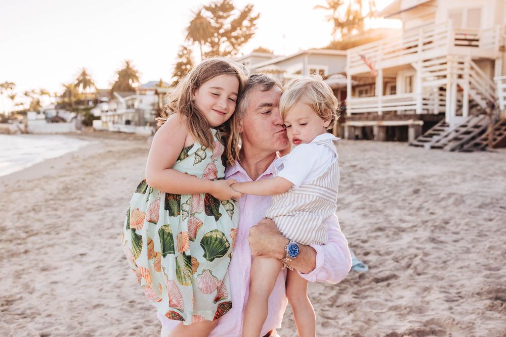Father holding two young children on a sunny beach, sharing a joyful moment at golden hour.