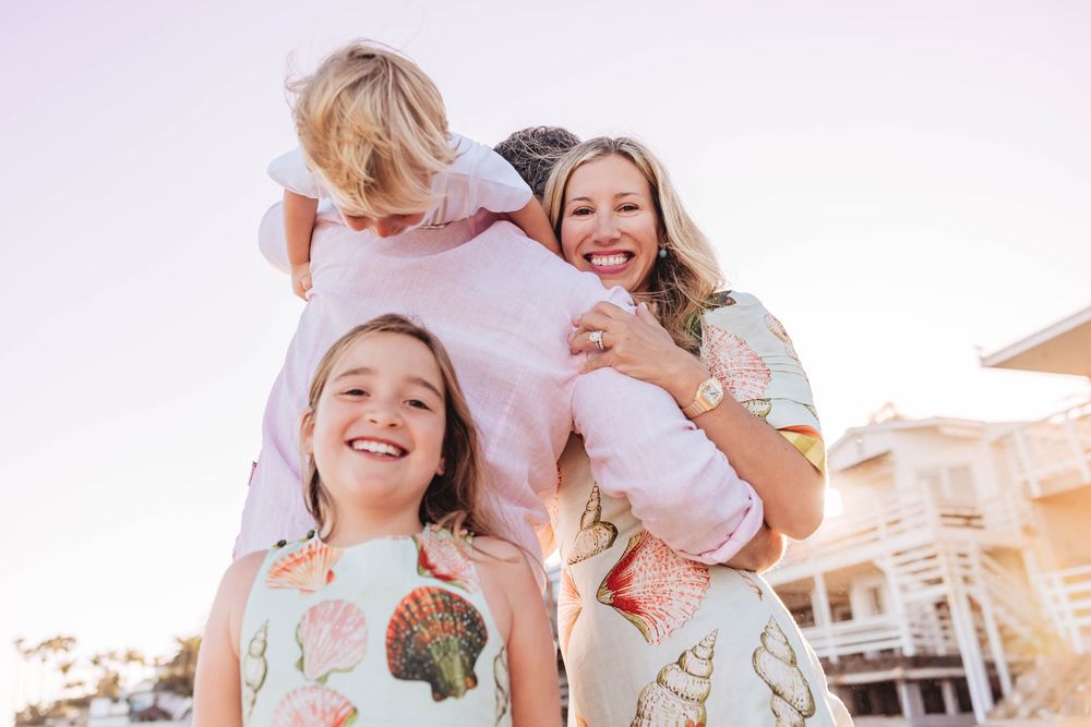 A smiling family of four poses outdoors, with a child on dad's shoulders near beachside homes.