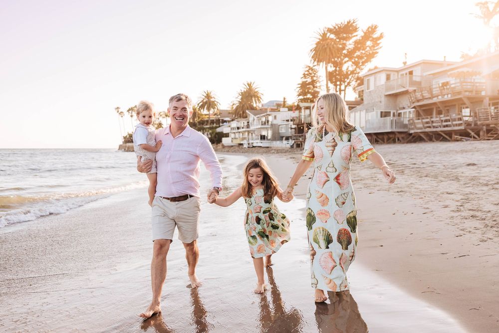 Family of four walking on a sunny beach, parents holding children's hands with coastal homes in the background.