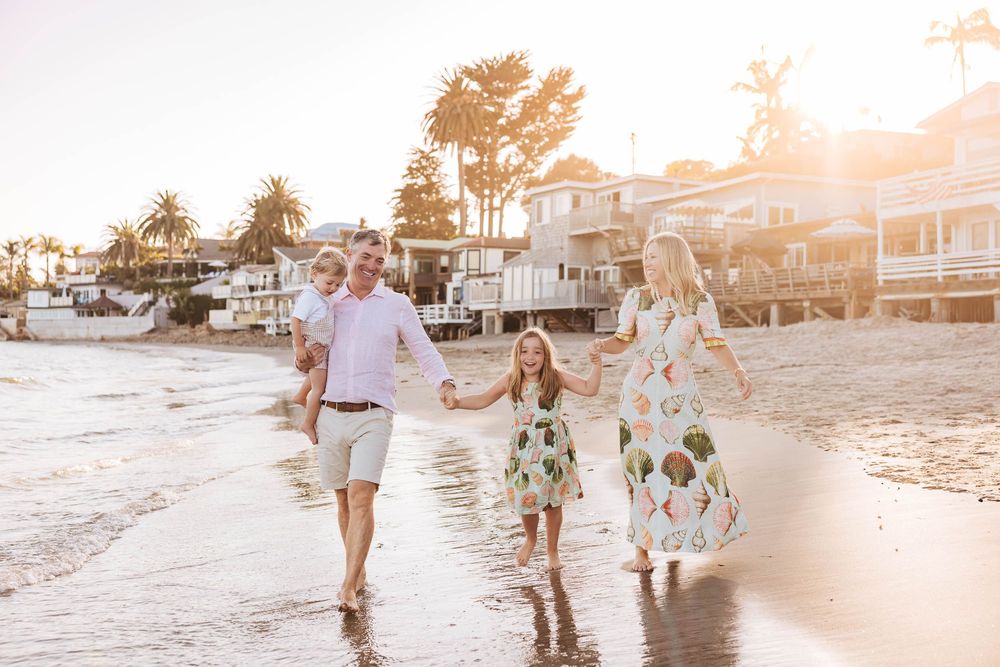 Family of four walking hand-in-hand on a sunny beach at golden hour near coastal homes.