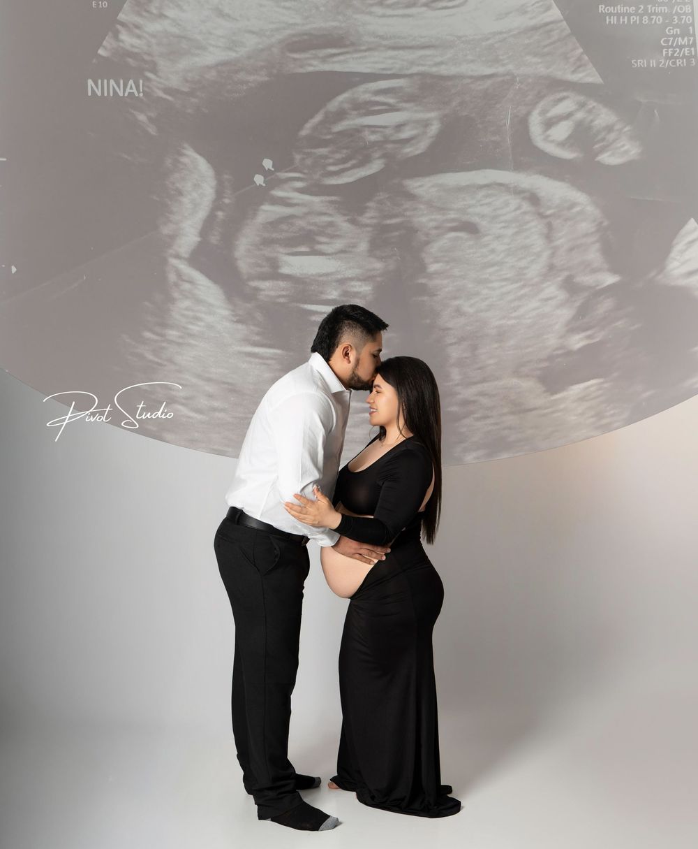 Couple in maternity photo shoot embracing before ultrasound backdrop, woman in black dress, man in white shirt.