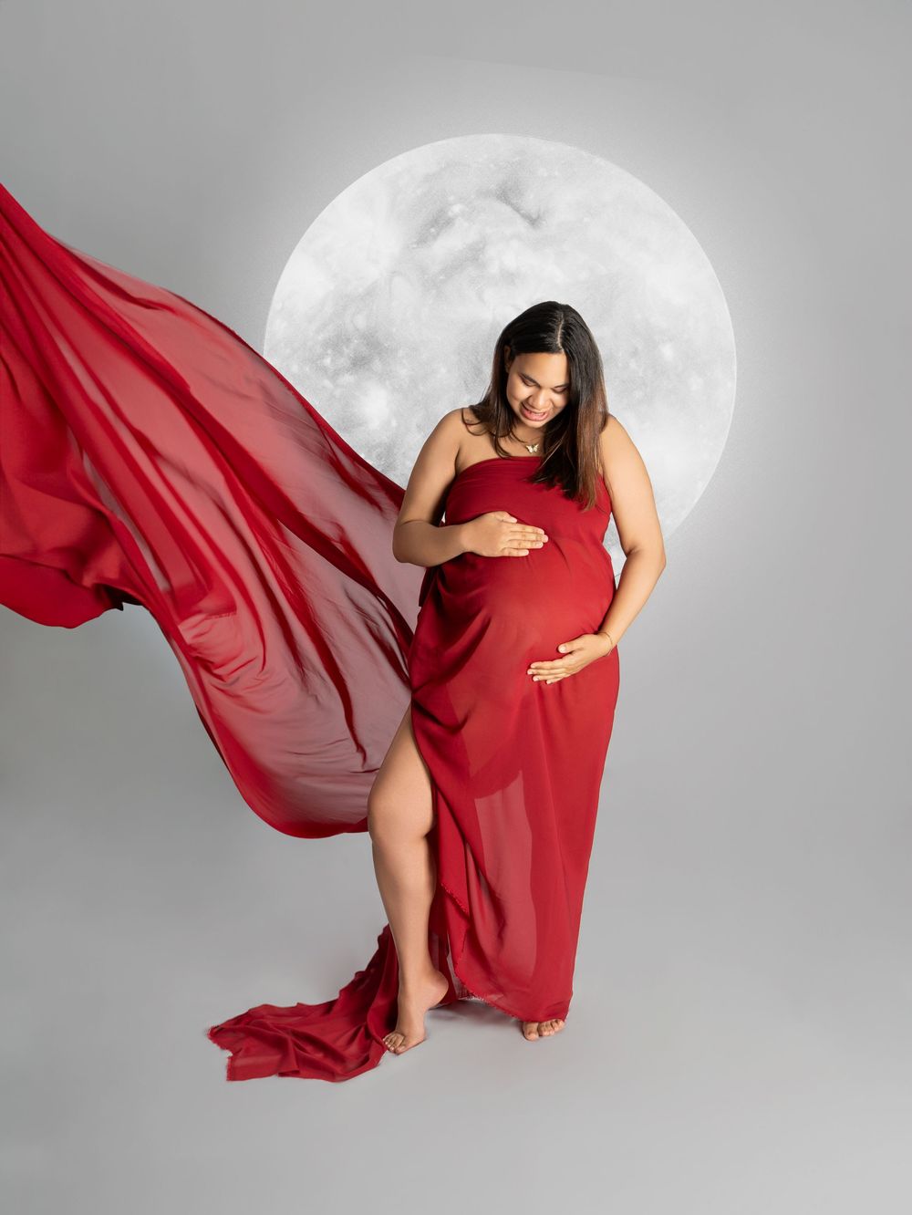 Pregnant woman in flowing red gown poses gracefully before a full moon backdrop in a maternity photo session.