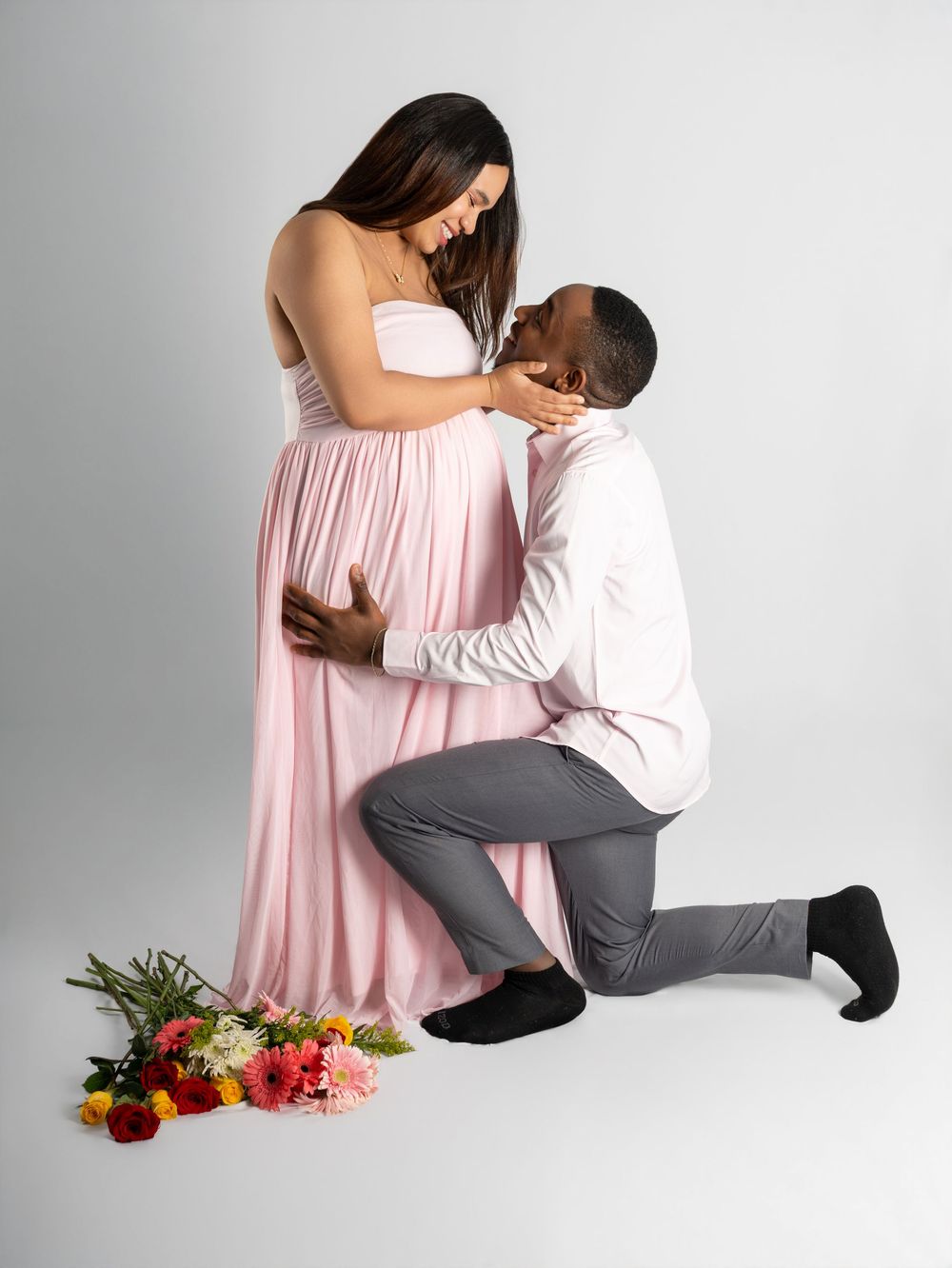 Pregnant woman in pink gown with partner kneeling beside her, surrounded by colorful flowers in a studio maternity shoot.