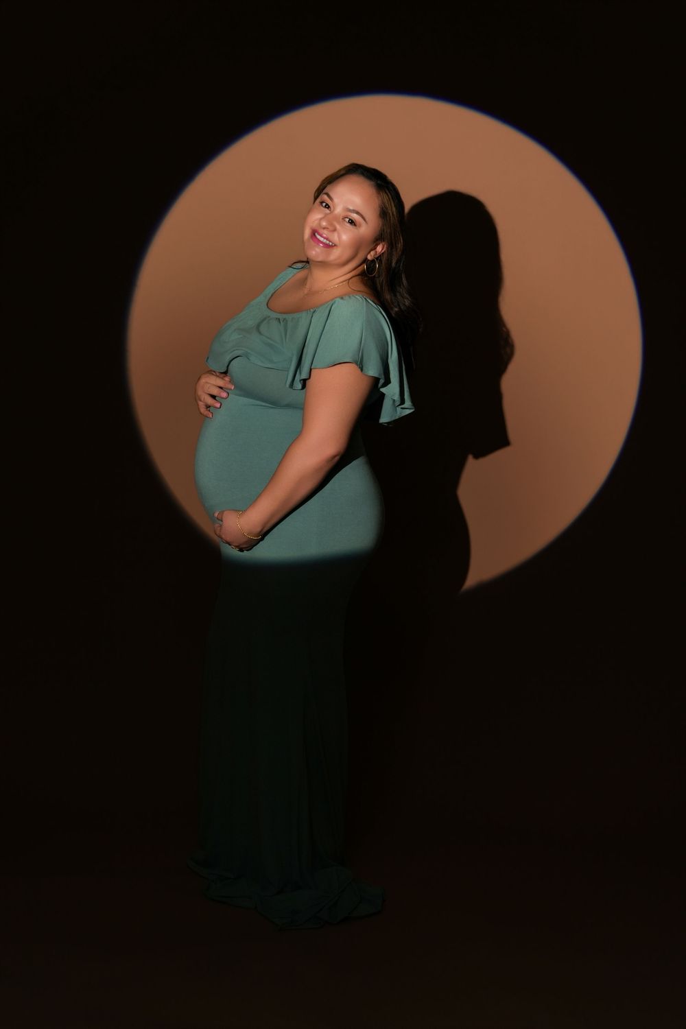 Pregnant woman in teal dress poses in circular spotlight against dark background, cradling baby bump.