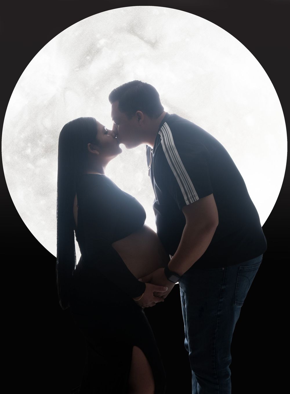 Couple sharing a kiss in silhouette against a glowing full moon backdrop, showcasing a beautiful maternity photo shoot.