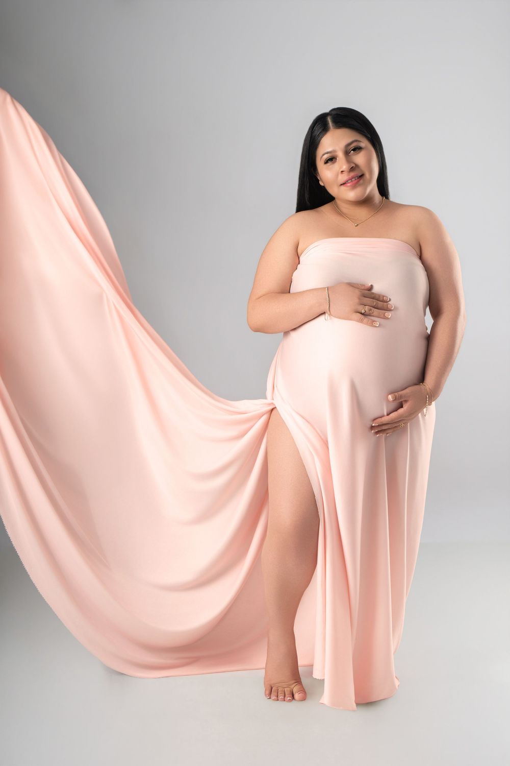 Pregnant woman in flowing pink maternity gown cradling her baby bump against a soft gray studio background.