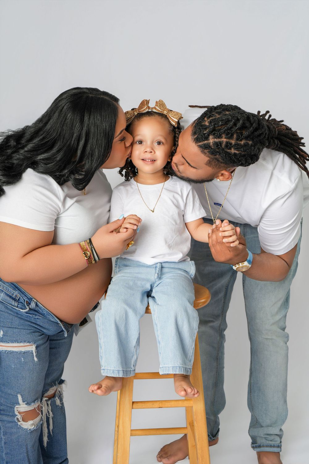 Pregnant woman and man kissing cheeks of toddler girl in gold bow sitting on stool, family maternity photo.