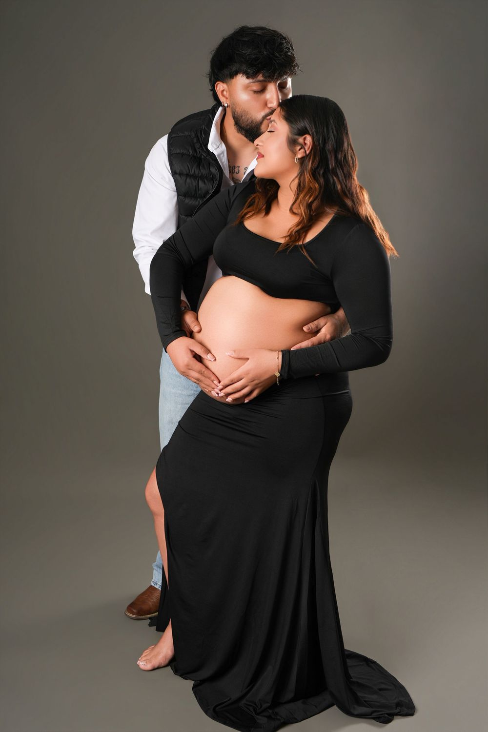 Couple sharing a tender kiss during a maternity photoshoot, woman in black dress cradling her baby bump.