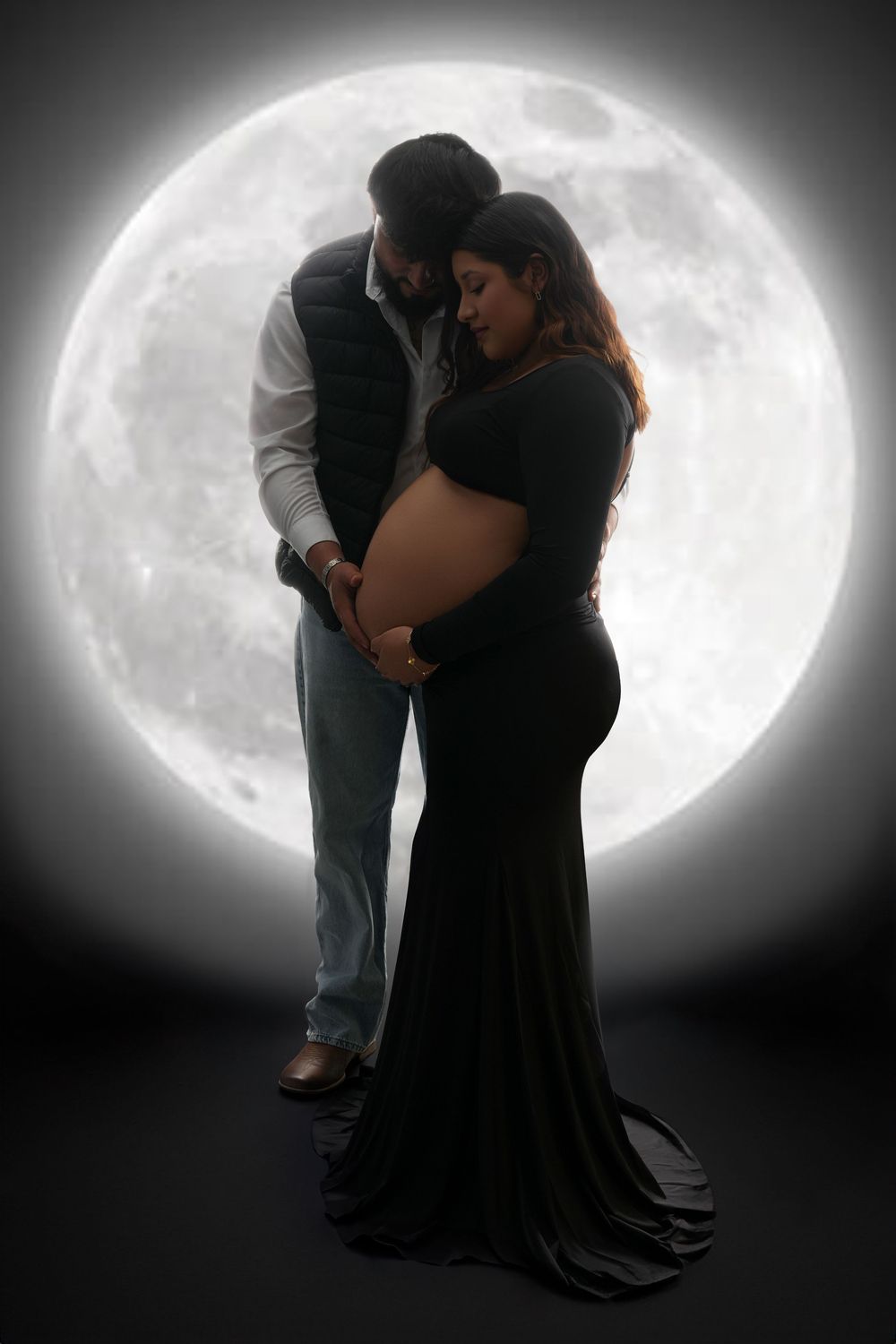 Couple in maternity photo shoot posed before glowing full moon, woman in black gown cradling baby bump.