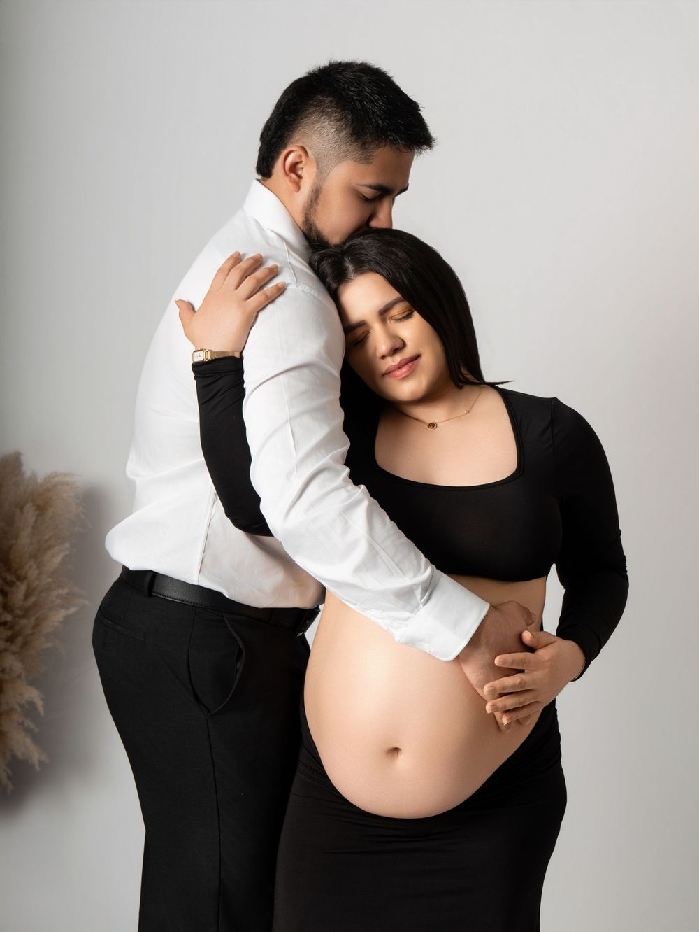 Couple embracing during maternity photoshoot, pregnant woman in black outfit with exposed belly, man in white shirt.