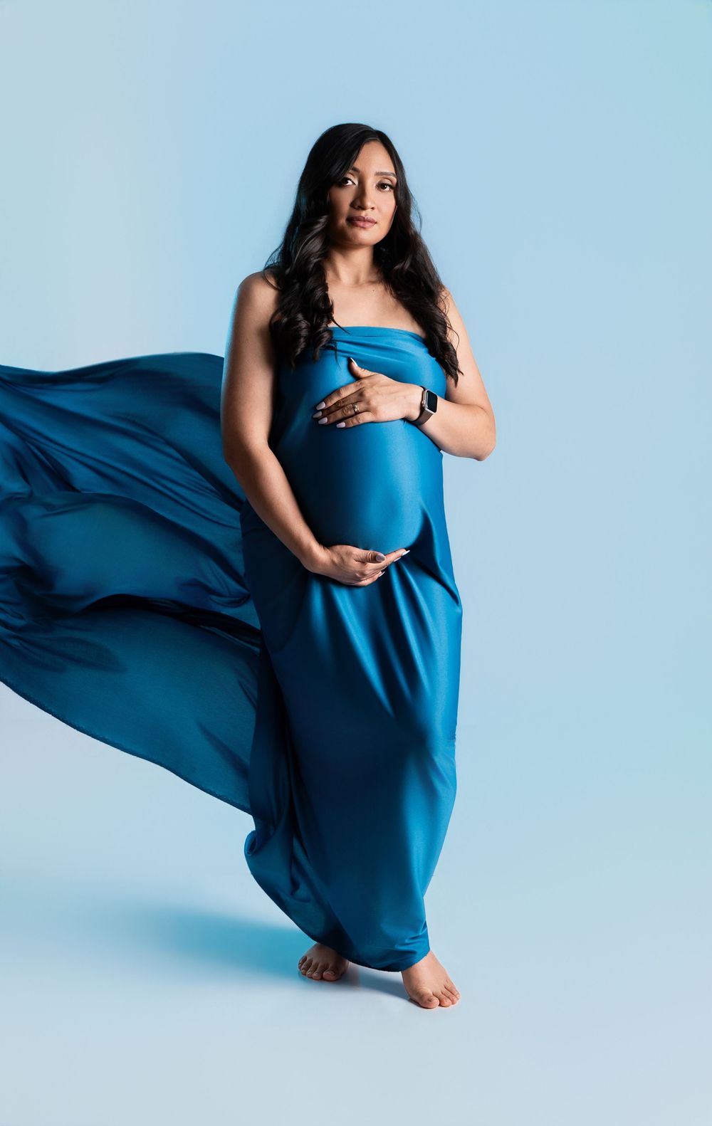 Pregnant woman in flowing teal silk dress cradles baby bump against light blue studio background.