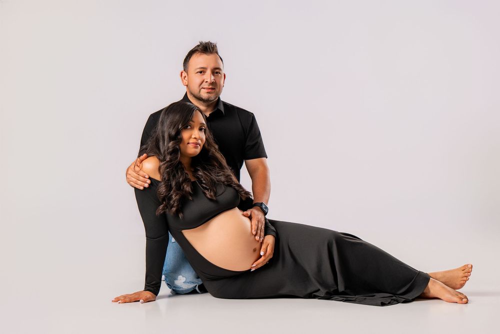Pregnant woman in black outfit sitting on floor with partner behind her in a professional maternity photo shoot.