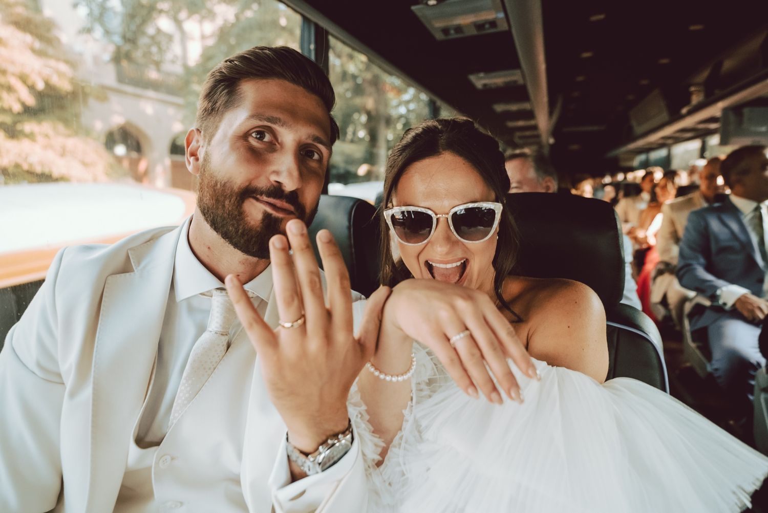 Newlyweds showing off wedding rings on a bus, bride in white gown and sunglasses, groom in white suit.