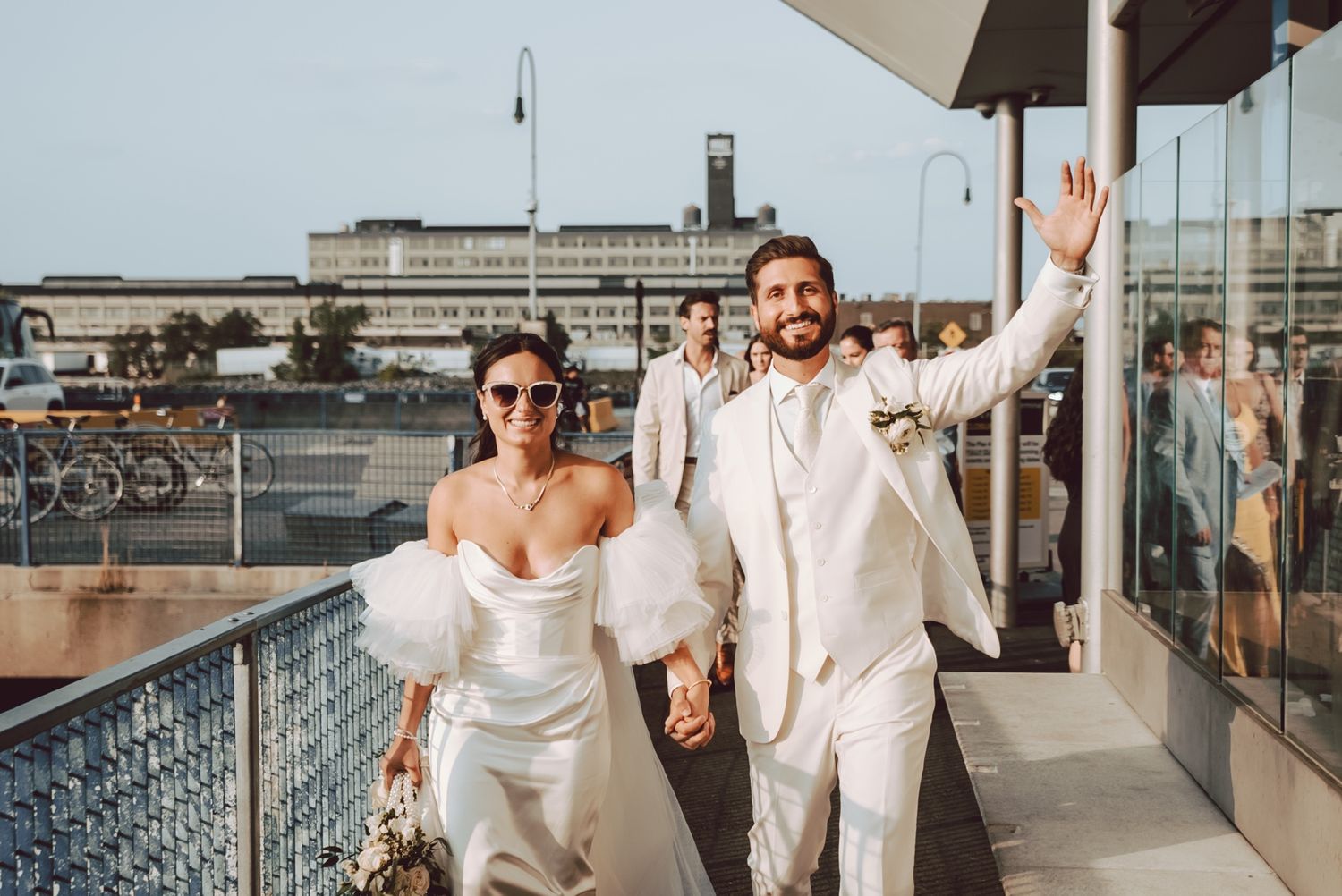 Joyful newlywed couple in white wedding attire walking on a waterfront venue balcony, groom waving.