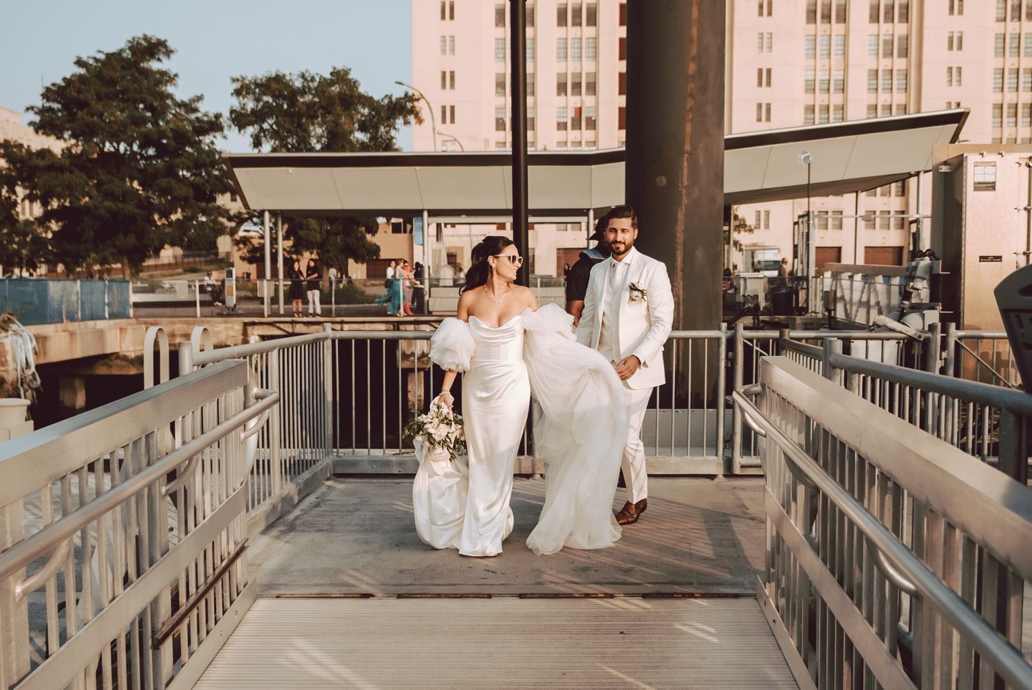 Bride and groom walk hand-in-hand on a waterfront bridge, bride in white gown, groom in white suit.