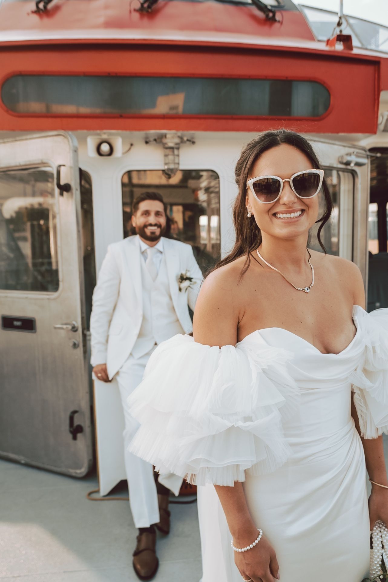 Smiling bride in off-shoulder white gown and sunglasses poses near red vehicle, groom in white suit behind her.