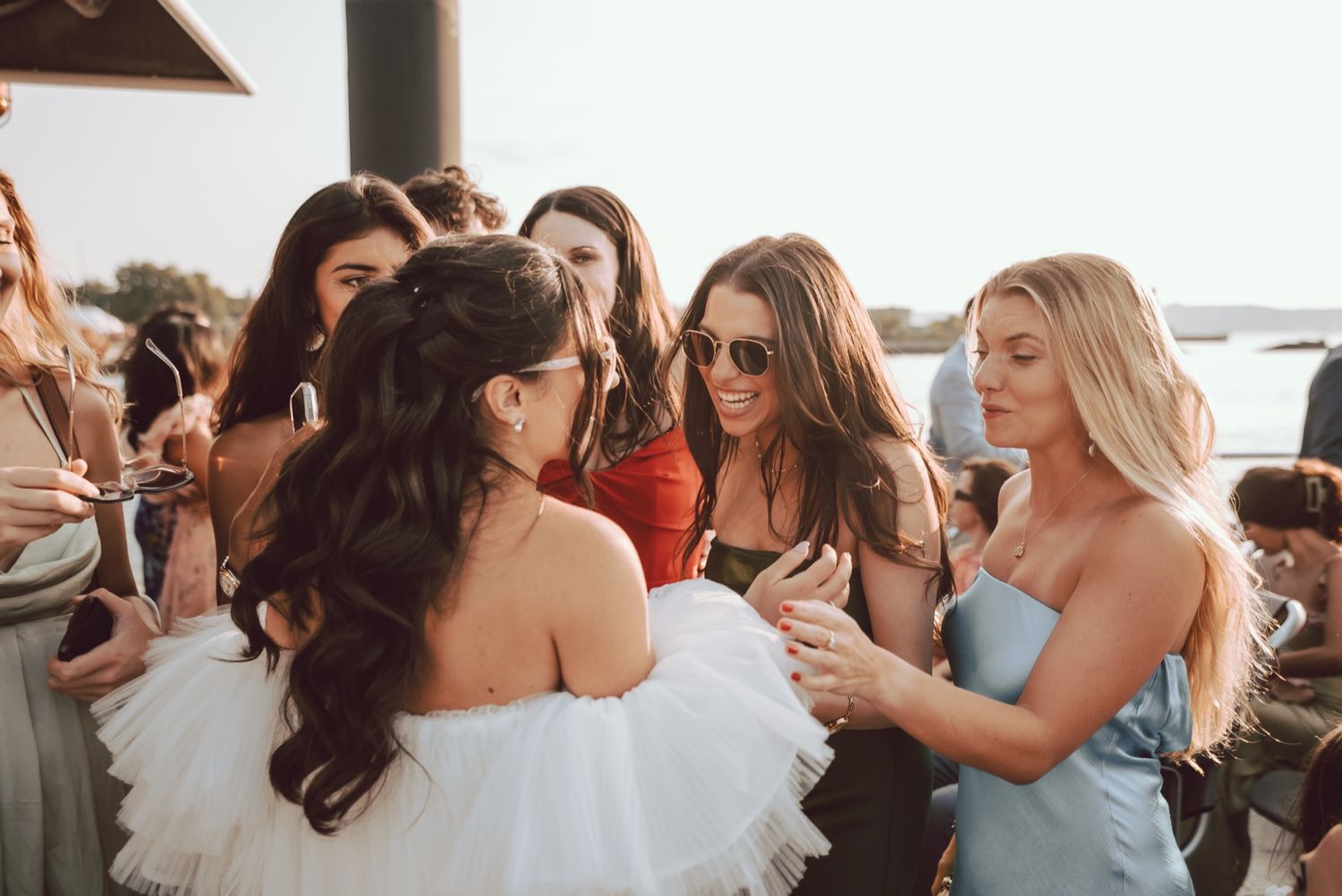 Bride in ruffled white gown celebrating with excited bridesmaids and guests at an outdoor wedding reception.