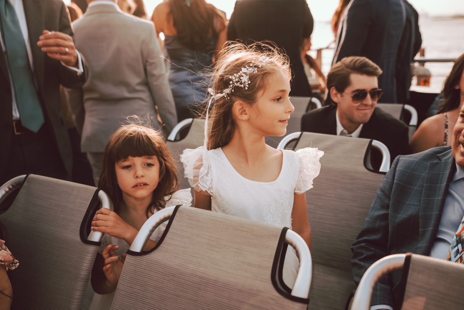 Two young flower girls in white dresses seated among wedding guests during an outdoor boat ceremony.