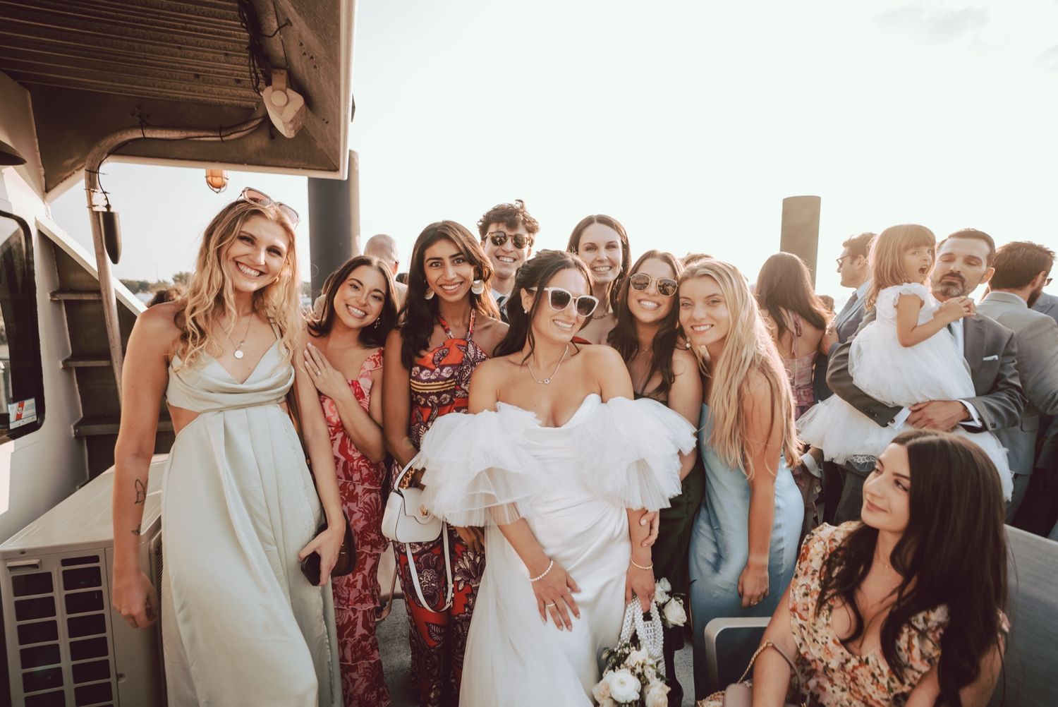 Bride in white dress poses with bridesmaids and guests at outdoor wedding celebration.