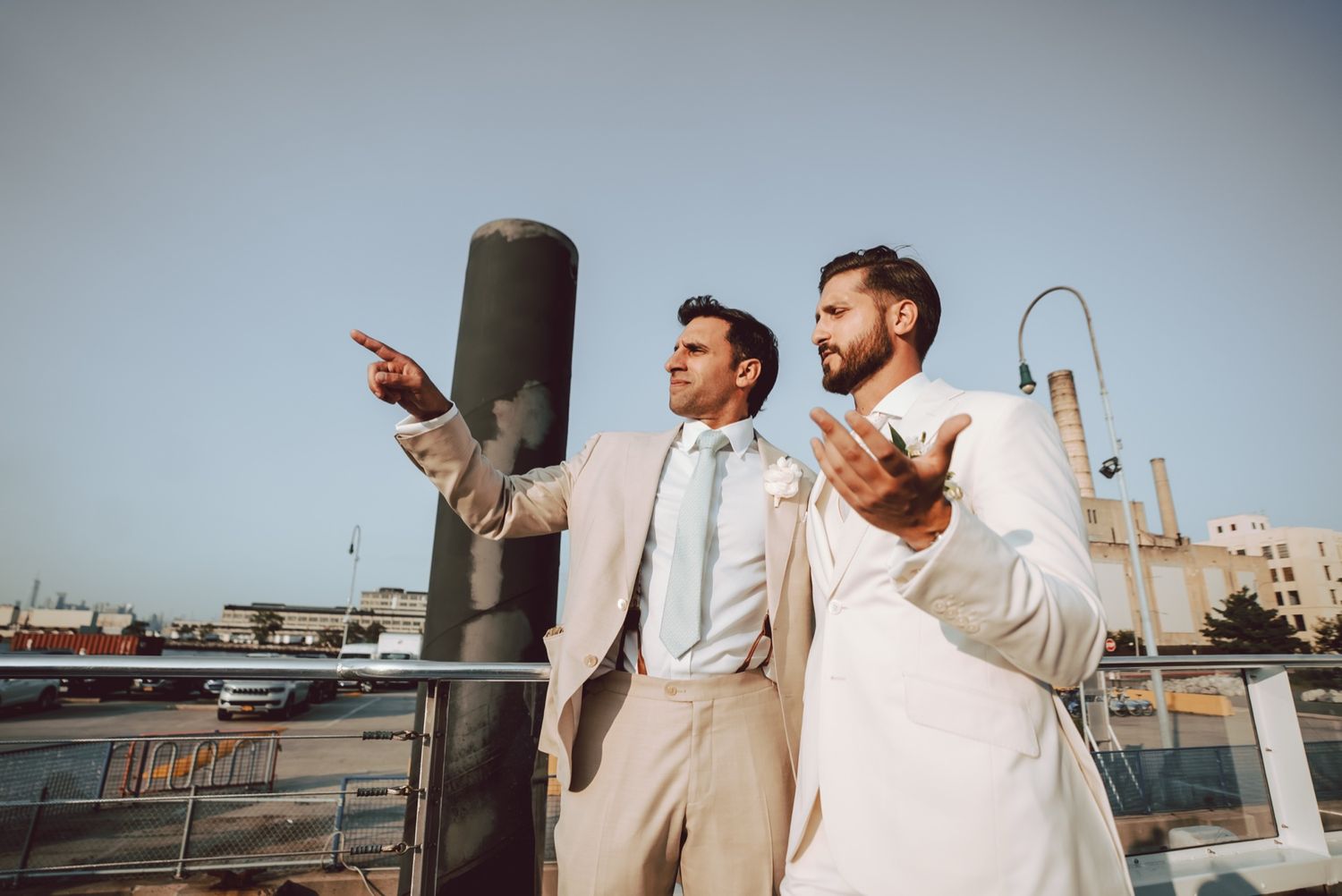 Two groomsmen in suits pointing and gesturing on a waterfront deck during a wedding celebration.