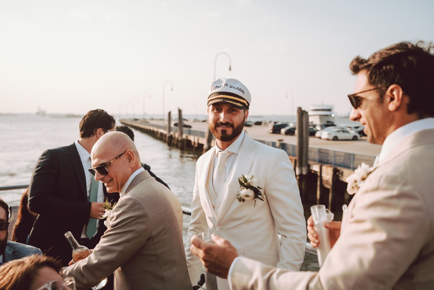 Groom in white suit and captain hat socializing with guests at an outdoor waterfront wedding reception.