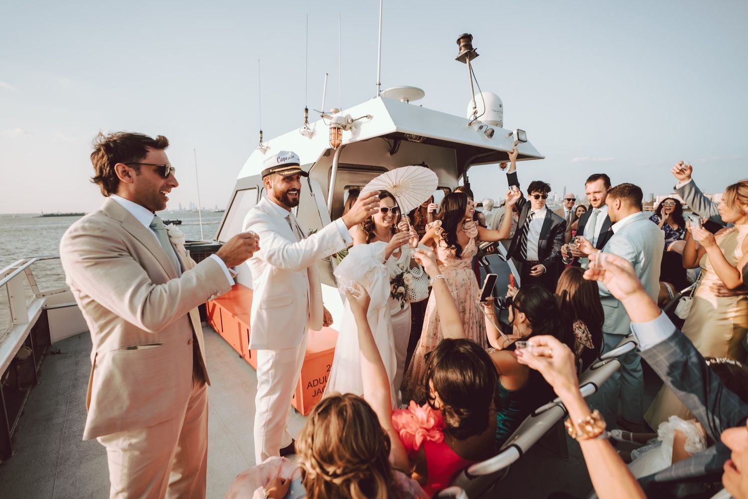 Bride and groom celebrating on a yacht with wedding guests, toasting champagne on a sunny day.