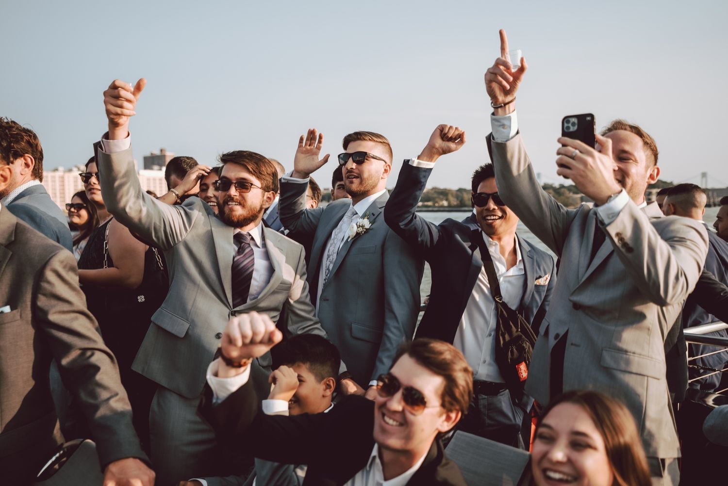 Groomsmen in gray suits celebrating outdoors, raising fists and taking selfies at a wedding reception.