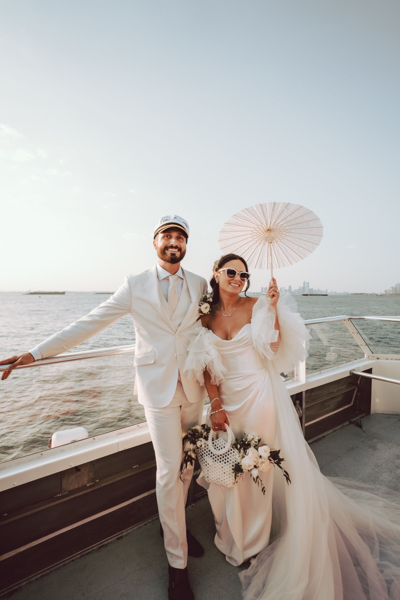 Bride and groom posing on a boat deck, bride holding white parasol and bouquet, groom in white suit.