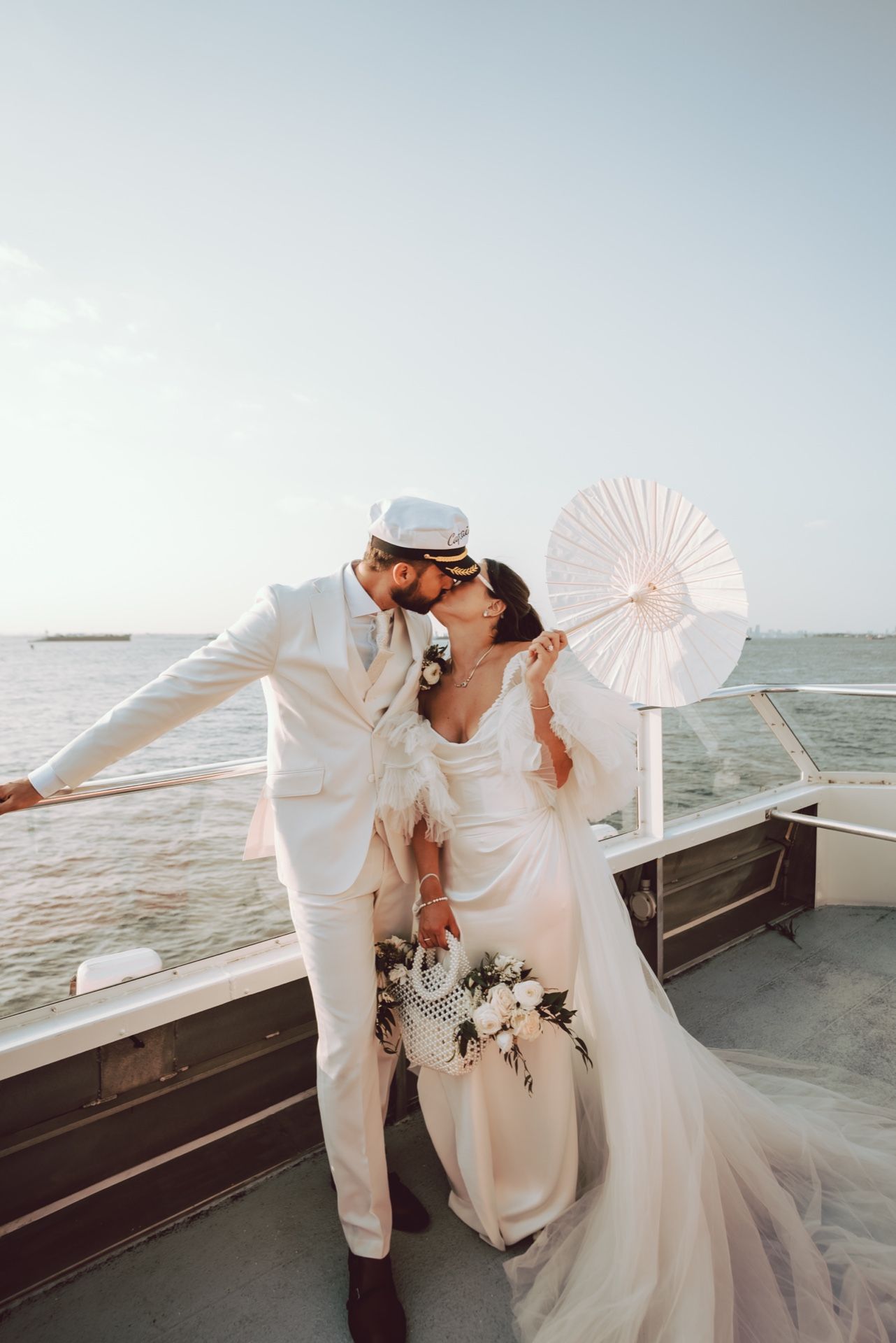 Bride and groom kissing on boat deck, bride holding white fan and bouquet, ocean backdrop.