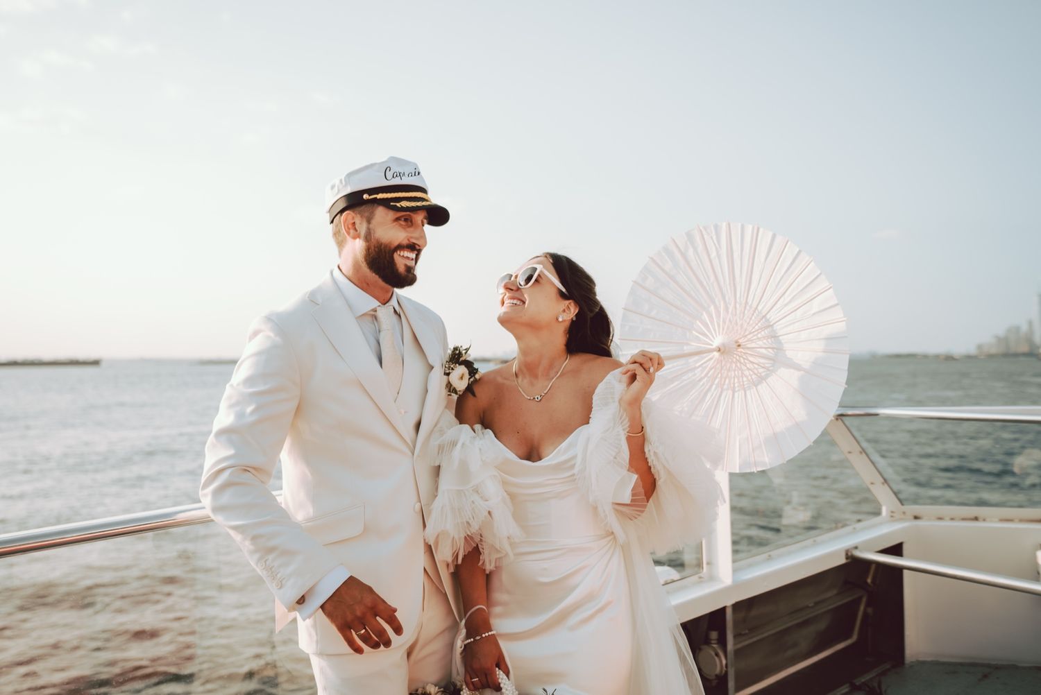 Bride and groom in white attire smiling on a boat deck, bride holding a white paper fan parasol.