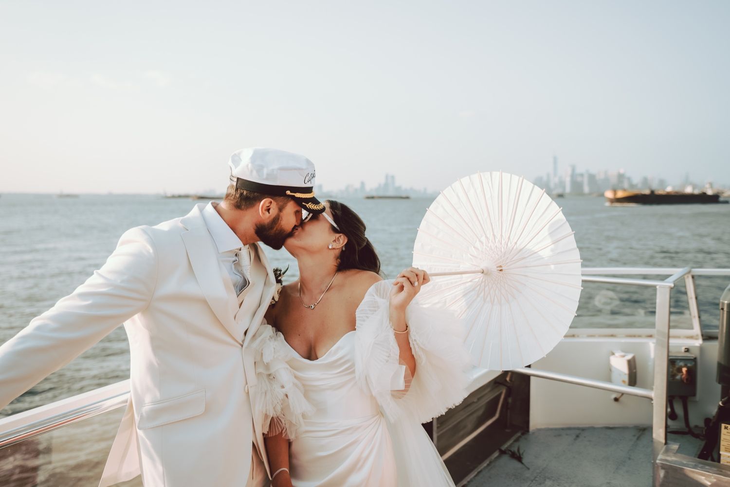 Newlyweds sharing a kiss on a yacht with city skyline and ocean in the background.