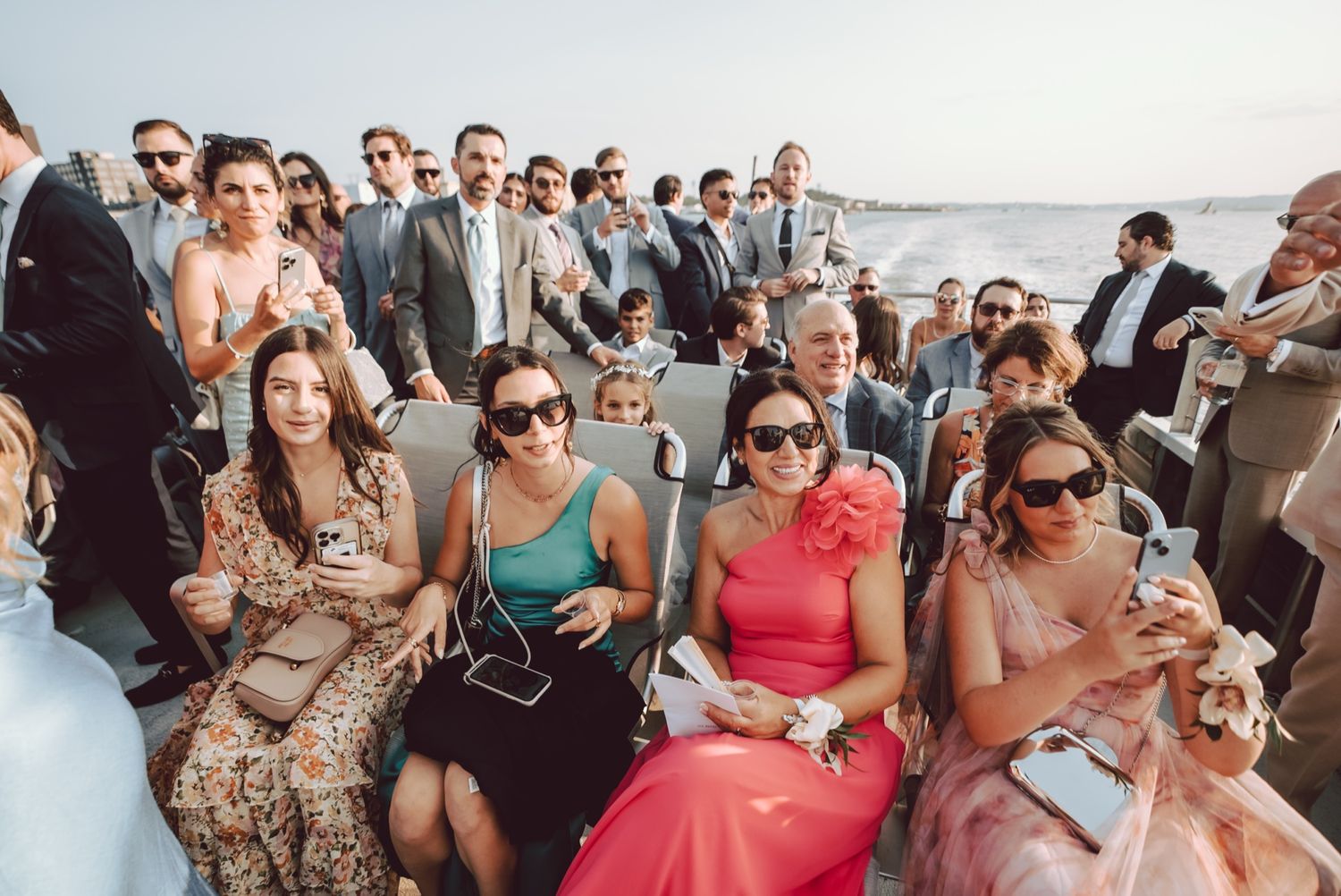 Wedding guests seated and standing on a boat deck, dressed in colorful formal attire outdoors.