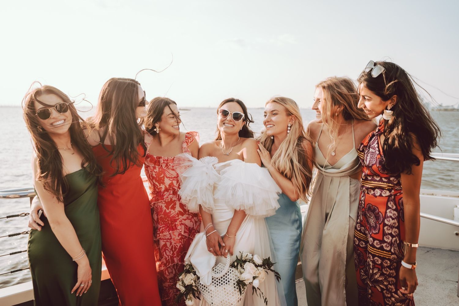 Bride in white ruffled dress laughing with six bridesmaids in colorful gowns on a boat.